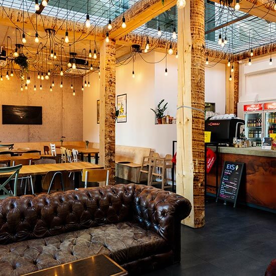 Gemütlicher Innenbereich im Stuntwerk mit vielen Sitzgelegenheiten (Holztische, Ledersofa). An der Decke hängen viele Glühbirnen, die den Raum in warmes Licht tauchen. Rechts ist die Theke, dort steht eine große Kaffeemaschine und zwei Getränkekühlschränke. Eine handbeschriebene Tafel wirbt für Eis.