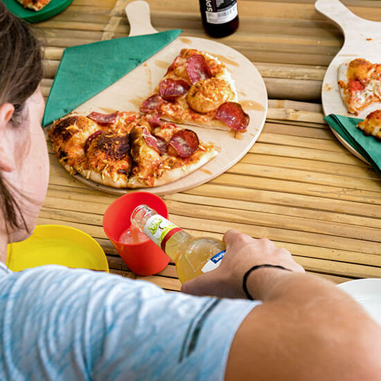 Kinder sitzen gemeinsam am Tisch und essen Pizza. Ein Kind schenkt Apfelschorle in bunte Becher ein