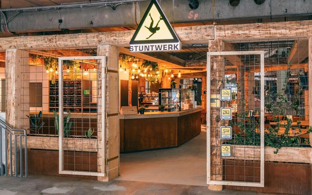 Eingangsbereich des Stuntwerk Troisdorf mit markantem Holzrahmen, Pflanzen und beleuchtetem Logo-Schild. Einladender Startpunkt in der Boulder- und Fitnesswelt.