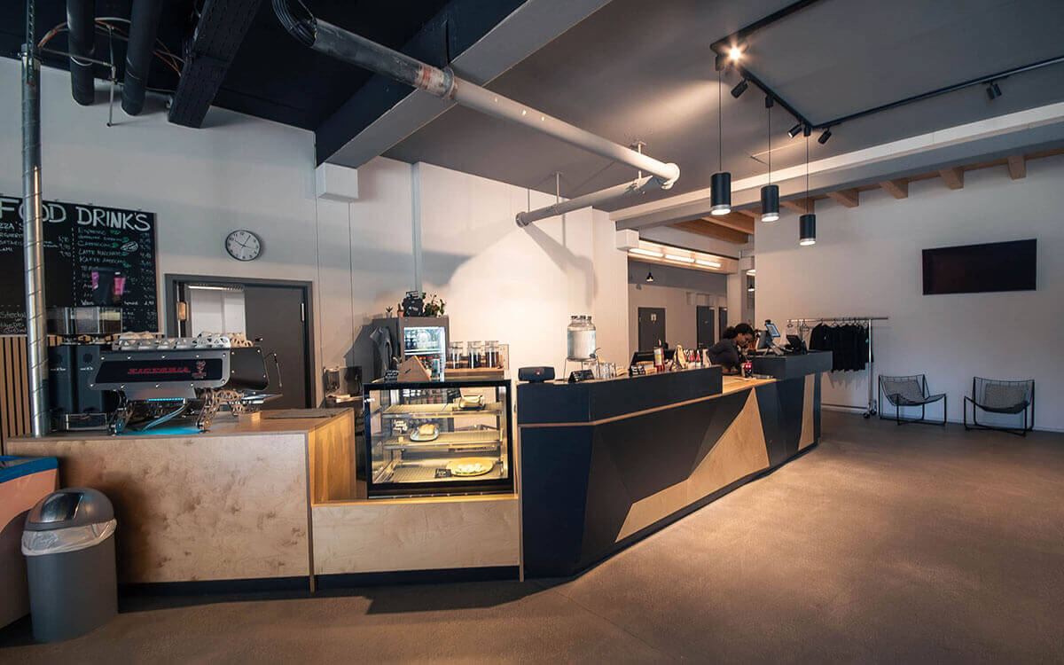 Moderner Empfangsbereich mit geometrischer Holz- und Schwarztresen-Theke, Glasvitrine mit Kuchen und industriellem Decken-Design im Stuntwerk Trostberg.