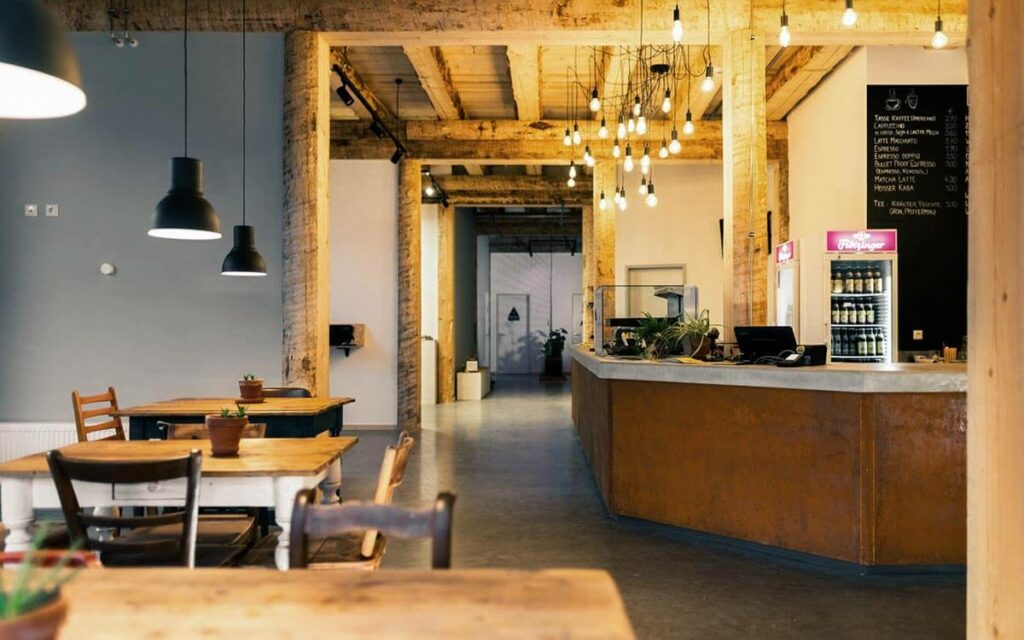 Eingangsbereich mit Blick auf den Check-In und das Bistro. Glühbirnen an der Decke tauchen die Szene in ein weiches Licht. Rustikale Tische und Stühle laden zum Verweilen ein. Grünpflanzen sorgen für eine Wohnzimmeratmosphäre.