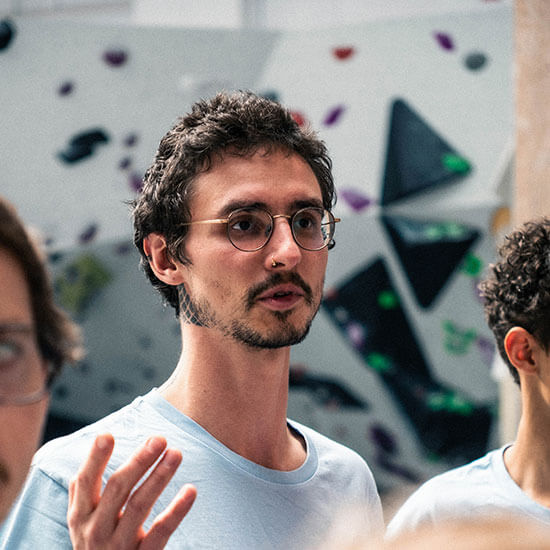 Junger Mann mit Brille spricht in einer Boulderhalle und gestikuliert leicht.