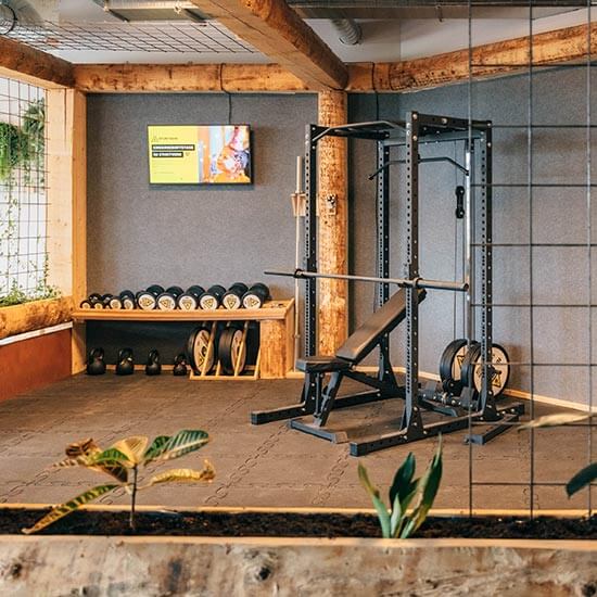 Gemütlich gestalteter Trainingsbereich mit Holzbalken, Pflanzen, grauen Wänden und einer Hantelbank. Kurzhanteln, Kettlebells und Stuntwerk-Beschilderung runden das warme Design ab.