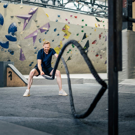 Sportler trainiert mit Battle Ropes vor einer bunten Boulderwand mit gelben, grünen und violetten Griffen im Stuntwerk. Intensives Functional Training in energiegeladener Atmosphäre.