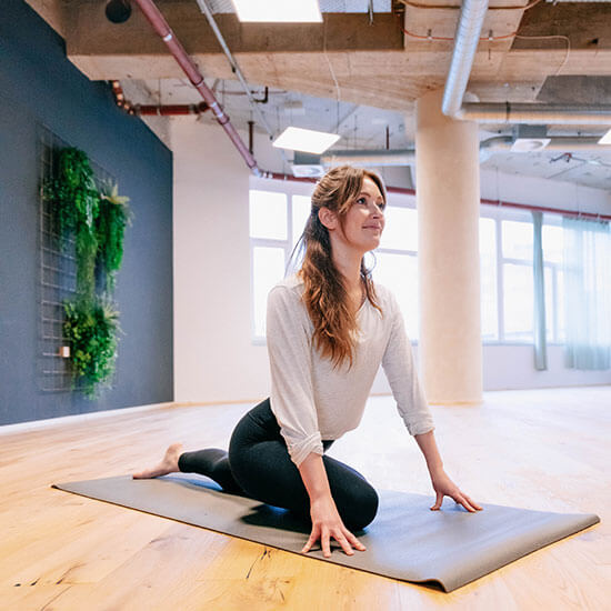 „Frau praktiziert die Schwan-Pose auf einer Yogamatte im Stuntwerk. Großes Fensterlicht und warme Holztöne verleihen der Szene eine ruhige, offene Stimmung.