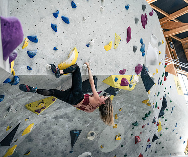 Eine sportliche Frau bouldert an einem Überhang in der Halle des Stuntwerk Rosenheim. Ihre Haare wehen nach unten, sie hält sich an den bunten Bouldergriffen fest. An der grauen Boulderwand sind viele bunte Routen verschiedener Schwierigkeitsstufen gebaut. Im Hintergrund oben erkennt man die Holzdecke.