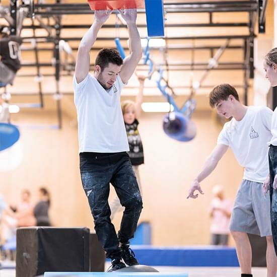 Mehrere Teilnehmende trainieren gemeinsam im Ninja-Bereich des Stuntwerks. Eine Person balanciert über ein Hindernis, während andere unterstützen und Hinweise zur Technik geben.