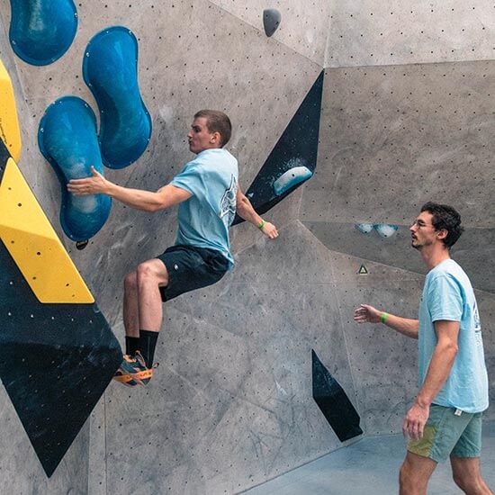 Ein junger Mann klettert an einer grauen Boulderwand mit großen blauen Volumen. Ein zweiter Mann steht hinter ihm um ihn zu supporten.