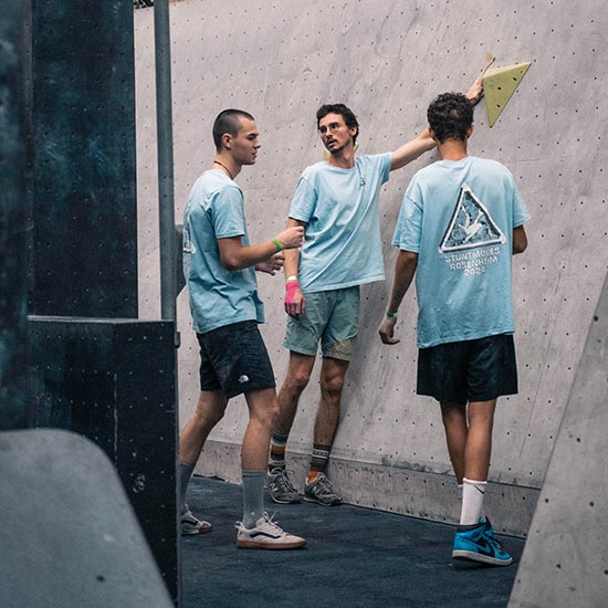 Drei Bouldernde in hellblauen Shirts besprechen gemeinsam eine Route an der grauen Boulderwand.