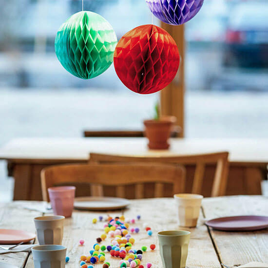 Festlich gedeckter Geburtstagstisch im Stuntwerk mit bunten Hängekugeln, Papptellern und farbigen Pompons. Im Hintergrund große Fensterfront – perfekte Kulisse für eine Kinderparty.