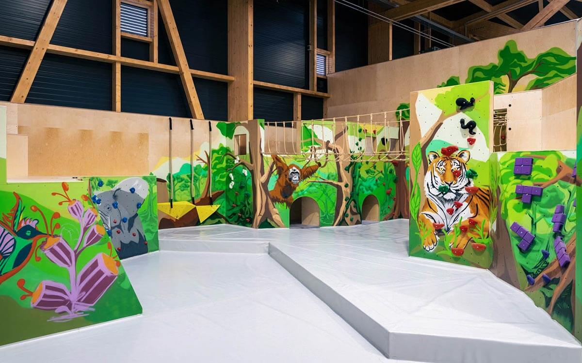 Kinderbereich mit weichen Fallschutzmatten für mehr Sicherheit. Eine Hängebrücke, kindgerechte Bouldergriffe und Seile laden Kinder zum Klettern ein. Die Wände sind mit Dschungelmotiven gestaltet.