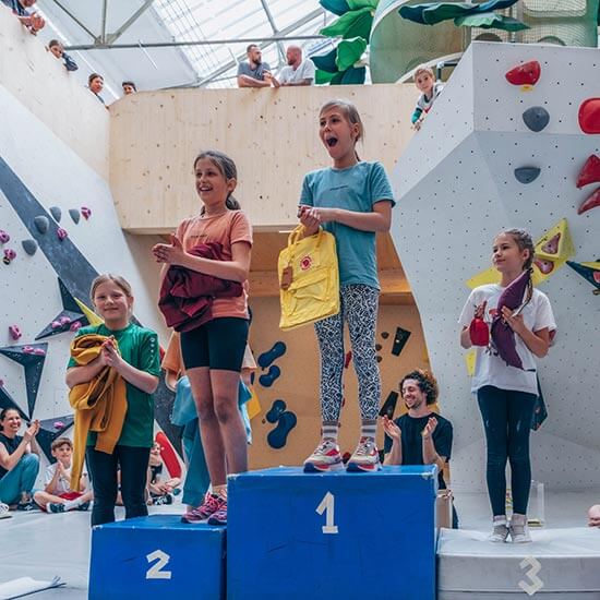 Kinder jubeln bei der Siegerehrung des Kidsmoves Wettkampfs im Stuntwerk. Drei junge Teilnehmerinnen stehen auf dem Podest und halten ihre Preise in den Händen.