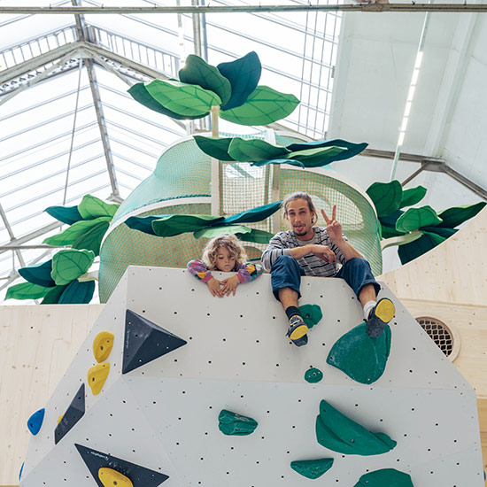 Ein Erwachsener und ein Kind sitzen gemeinsam oben auf einer weißen Boulderwand im Kinderbereich des Stuntwerks Köln Mülheim. Hinter ihnen ragt der grüne Kletterbaum mit großen Blättern bis unter das Glasdach – fröhlicher Moment voller Bewegung, Nähe und Abenteuer.