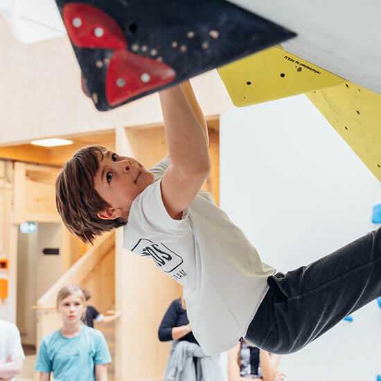 Kind klettert konzentriert an gelben Klettergriffen in der Boulderhalle des Stuntwerks. Mit Chalk-Tasche und Kletterschuhen arbeitet es sich Schritt für Schritt nach oben – Fokus, Technik und Spaß im Kids-Bouldern.