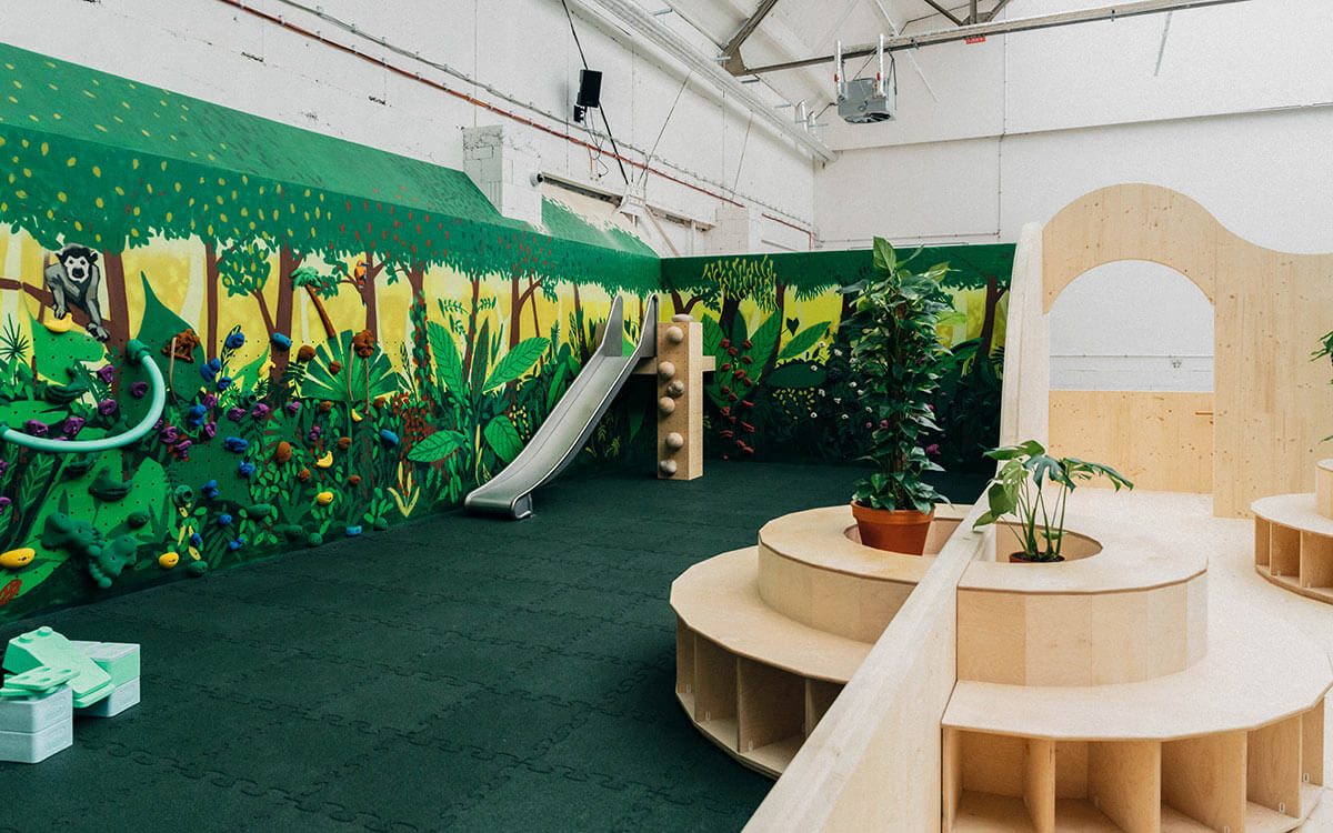 Farbenfroher Kinderbereich im Stuntwerk Köln Mülheim mit Dschungelwand, bunten Klettergriffen, Rutsche und Pflanzen. Die Wand zeigt tropische Blätter, Tiere und Bäume – ein kreativer Bewegungsraum für kleine Abenteurer:innen.