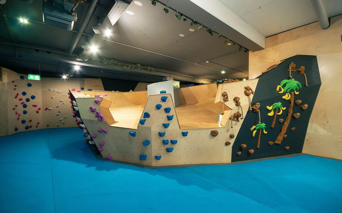 Verspielter Kinder-Kletterbereich mit blauen Matten, Holz- und Kletterwänden, bunten Griffen in Pink und Blau sowie dekorativen Palmen und Affenfiguren im Stuntwerk Trostberg.