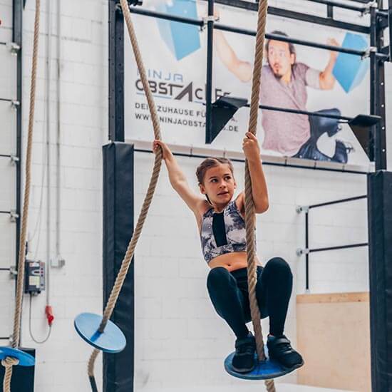 Mädchen klettert konzentriert an Seilen im Ninja-Parcours des Stuntwerks. Kraft, Gleichgewicht und Mut beim Kinder-Ninja-Training.