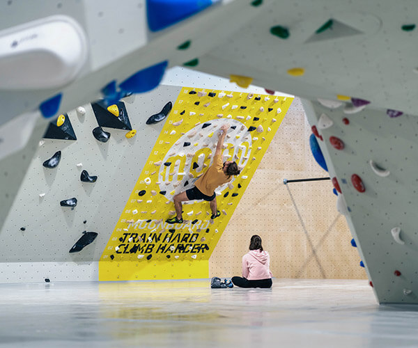 Blick von weiter entfernt unter einem Überhang hindurch auf das Moonboard. Das Moonboard ist gelb und leicht geneigt. Daran klettert ein Mann in Boulderschuhen und gelbem Shirt. Er greift gerade nach einem Griff weiter oben. Auf dem Boden sitzt eine Frau im rosa Pulli und sieht ihm zu.