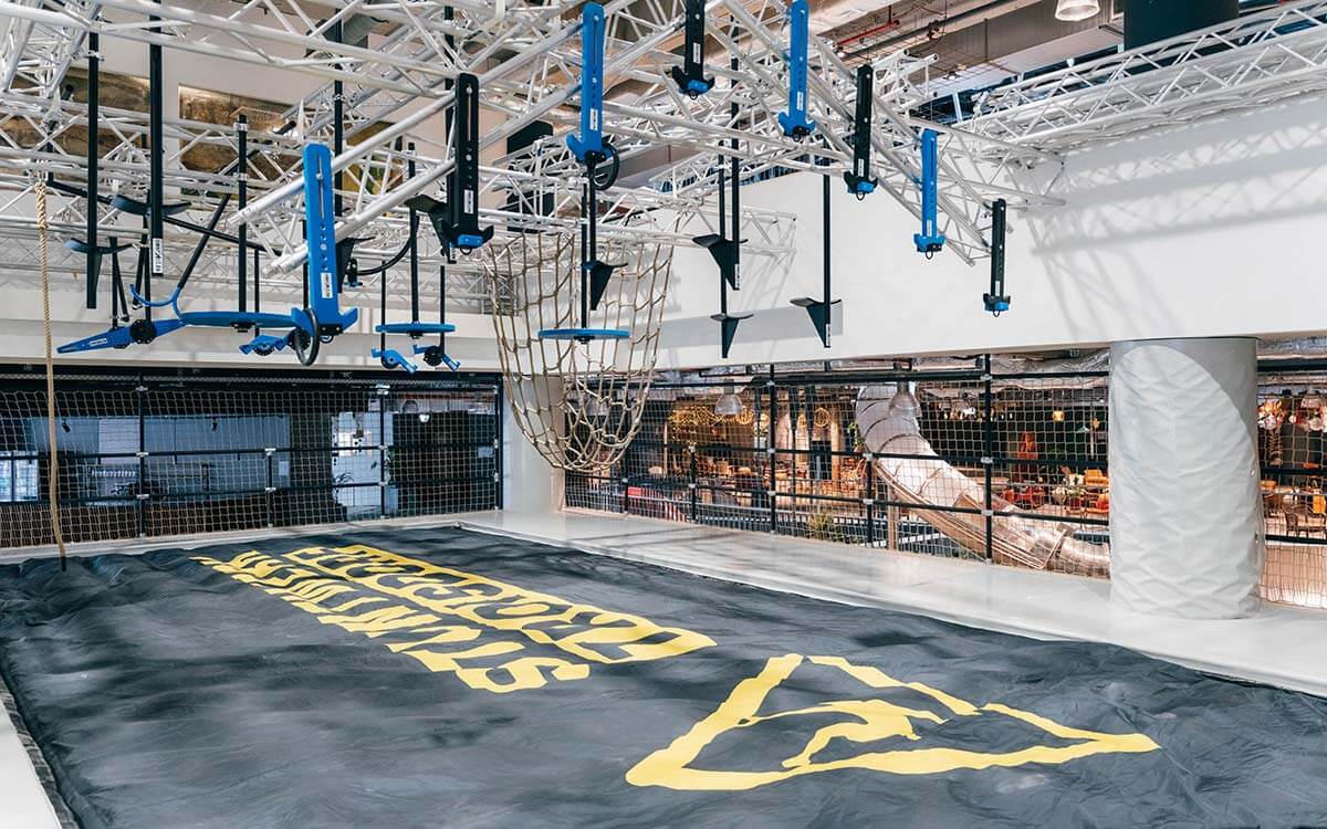 Moderner Ninja-Parcours im Stuntwerk Troisdorf mit blauen Hindernissen, Kletterseilen und Hangelstrecken aus Metall. Große schwarze Sicherheitsfläche mit gelbem Logo im Vordergrund.