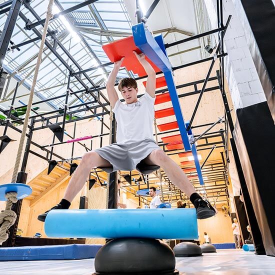 Jugendlicher trainiert im Ninja-Bereich des Stuntwerks an einem Balance- und Hang-Obstacle. Er hält sich an Griffen fest und balanciert über ein bewegliches Element in der Halle.