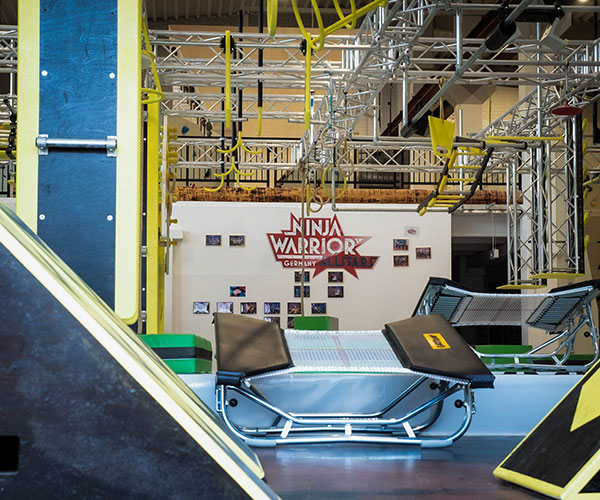 Ein großes Trampolin von dem Ninajframe. Im Ninjaframe hängen Hangelhindernisse wie Ringe und Stangen. Auf der Wand im Hintergrund hängt ein rotes Schild, auf dem "Ninja Warrior Germany Allstars" steht.