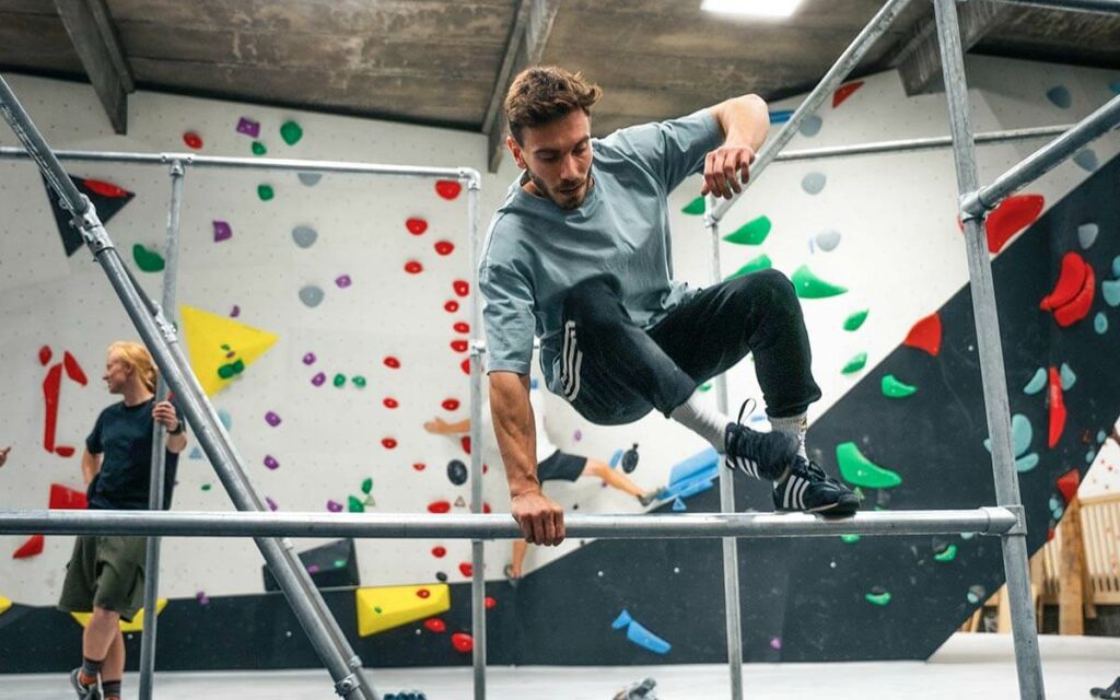 Person trainiert Parkour an einer Metallgerüst-Konstruktion in einer Halle mit bunten Boulderwänden; dynamische Bewegung beim Überspringen einer Stange, andere Personen trainieren im Hintergrund.