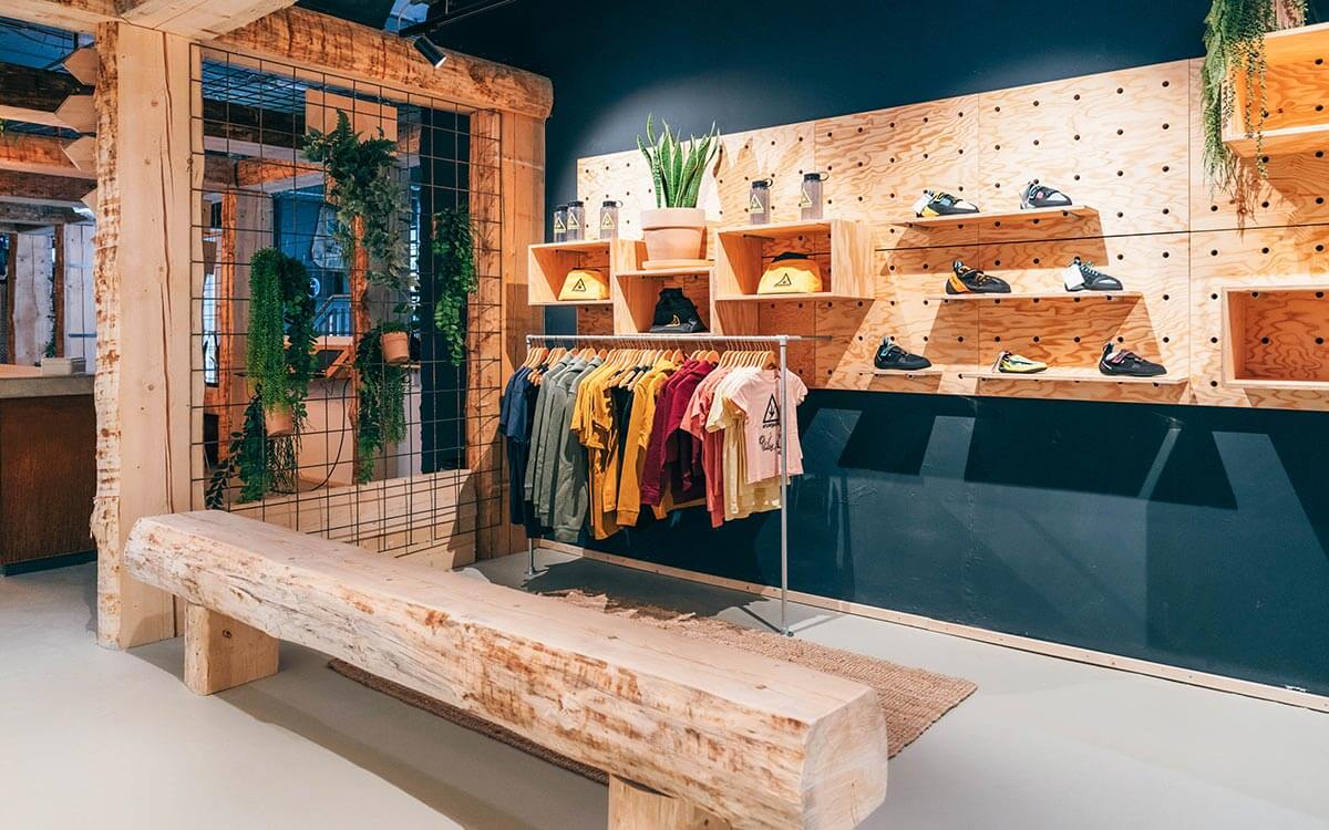 Stuntwerk-Shop in Troisdorf mit Holzregalen, grünen Pflanzen und Boulder-Merchandise. Bunt sortierte Shirts, Kletterschuhe und Accessoires in moderner, natürlicher Einrichtung.
