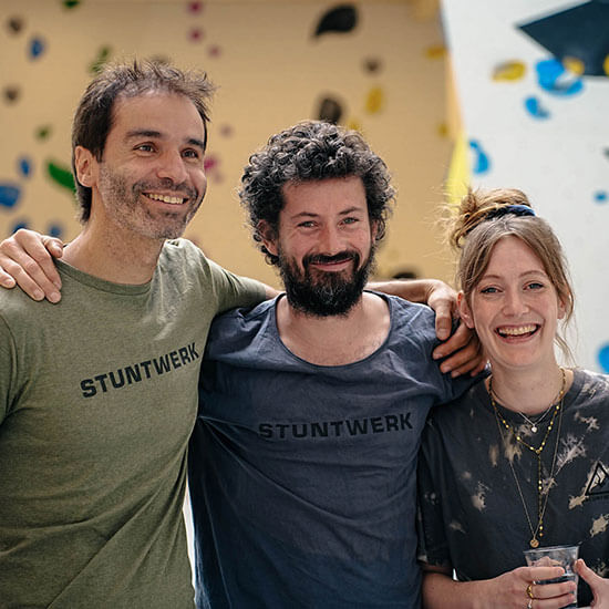 Drei Stuntwerk-Mitarbeitende (Uwe Pop, Florian Schiffer, Jana Jungbluth) stehen lachend vor einer Boulderwand. Sie alle tragen ein Stuntwerk Shirt in verschiedenen Farben.