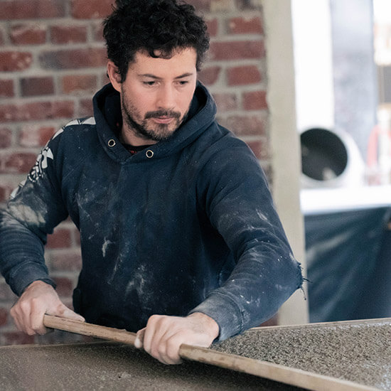 Florian Schiffer, Geschäftsführer der Stuntwerk GmbH, legt selbst Hand an. Er trägt einen blauschwarzen, mit Farbe und Dreck beschmierten Pullover. Er verteilt mit Hilfe eines Bretts Beton auf einer Oberfläche.