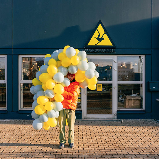 Im Hintergrund sieht man den Eingang des Stuntwerks Köln Zollstock. Das gelb-schwarze Logo hängt über der Glas-Eingangstür. Davor steht ein Mitarbeiter im Sonnenlicht. Es ist kalt, er trägt eine orange Mütze und orange Winterjacke. Er trägt eine große Traube an Luftballons in weiß-gelb, die ihn nahezu bedeckt.